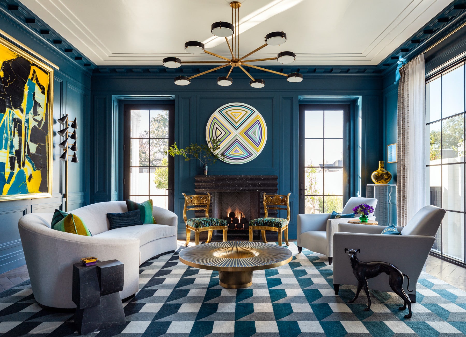 The Lounge features architectural paneling and a modillion cornice, providing a Neoclassical backdrop for vibrant colors and contemporary art and décor. A dramatic light fixture, echoing the sunburst motif introduced in the foyer, floats above the space, while Mid-Century-inspired sconces are integrated seamlessly into the richly painted wood paneling. A custom fireplace mantel carved from three blocks of Nero Marquina stone with an Art Deco motif, sits deeply in the wall, making an extension of the hearth beyond the fireplace surround unnecessary. Gilded antique chairs are paired with a modern curved sofa and vintage Donghia chairs. At the room’s center is a handcrafted brass and sodalite cocktail table and art piece by Belgian designer, Yann Dessauvages. (Photo Credit: Julie Soefer)